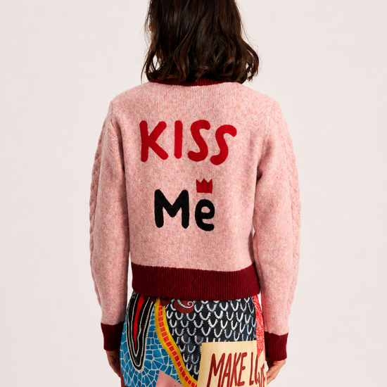 A model shown from the back wearing a light pink fuzzy cardigan with 'KISS Me' embroidered on the back in red and black, paired with a colorful patterned skirt against a plain off-white background.