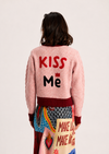 A model shown from the back wearing a light pink fuzzy cardigan with 'KISS Me' embroidered on the back in red and black, paired with a colorful patterned skirt against a plain off-white background.