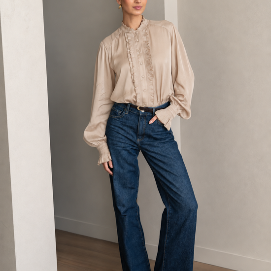 A woman stands in a minimalist room with light gray walls and wood floors, wearing a beige ruffled blouse and dark blue wide-leg jeans.