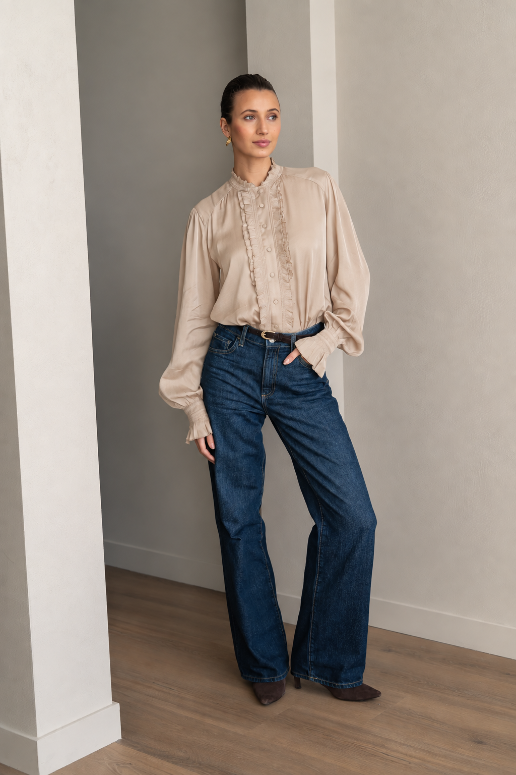 A woman stands in a minimalist room with light gray walls and wood floors, wearing a beige ruffled blouse and dark blue wide-leg jeans.