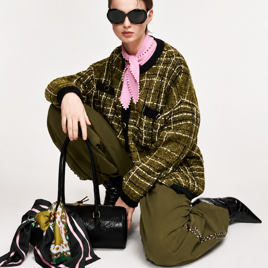 A model crouches on a light gray studio background wearing a green-and-black plaid jacket, olive green pants, a pink neck scarf, black boots, and black sunglasses. A black handbag with a colorful patterned scarf draped over it sits beside her.
