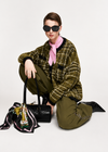 A model crouches on a light gray studio background wearing a green-and-black plaid jacket, olive green pants, a pink neck scarf, black boots, and black sunglasses. A black handbag with a colorful patterned scarf draped over it sits beside her.