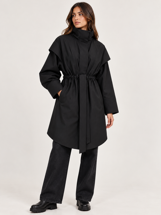 model wearing A black long-sleeve coat with a high mock neck, wide shoulder panels, and a self-tie belt at the waist, shown against a plain white background.