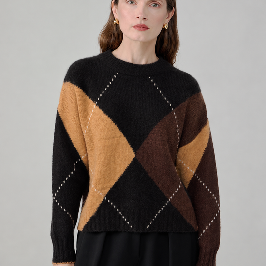 A woman modeling a black knit sweater with a large argyle pattern in tan, brown, and dark brown, paired with black trousers, against a neutral gray background.