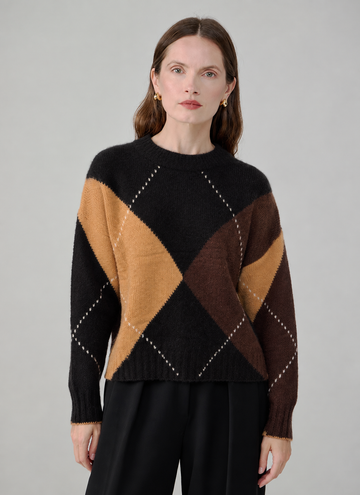 A woman modeling a black knit sweater with a large argyle pattern in tan, brown, and dark brown, paired with black trousers, against a neutral gray background.