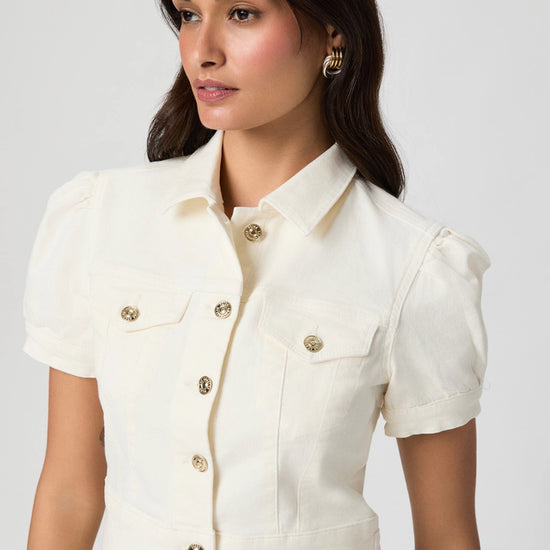 Woman wearing a white denim jacket with gold buttons on a plain background