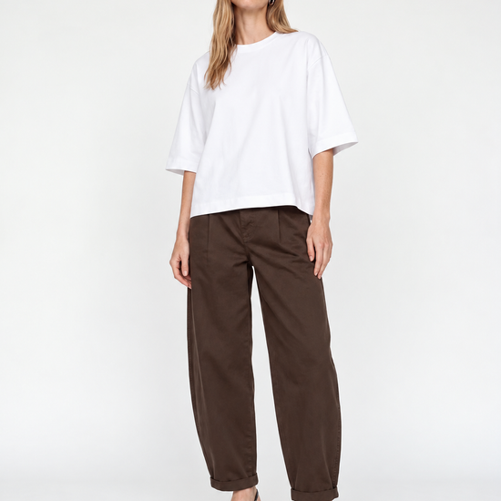 A woman modeling a white oversized short-sleeve top and dark brown wide-leg cropped pants with black pointed-toe heels, photographed against a clean white studio background.