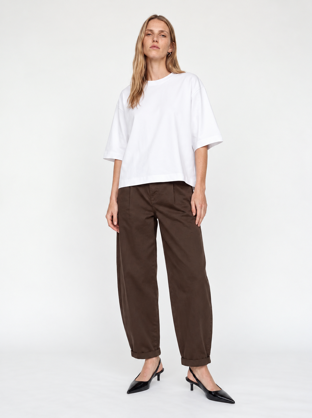 A woman modeling a white oversized short-sleeve top and dark brown wide-leg cropped pants with black pointed-toe heels, photographed against a clean white studio background.