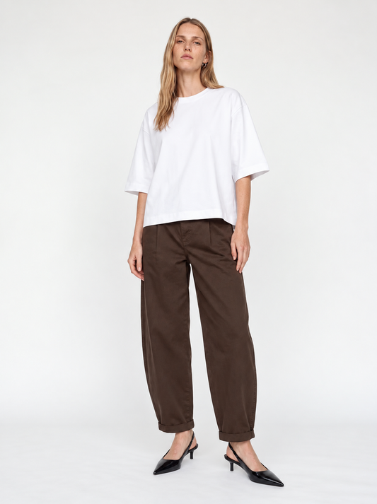 A woman modeling a white oversized short-sleeve top and dark brown wide-leg cropped pants with black pointed-toe heels, photographed against a clean white studio background.