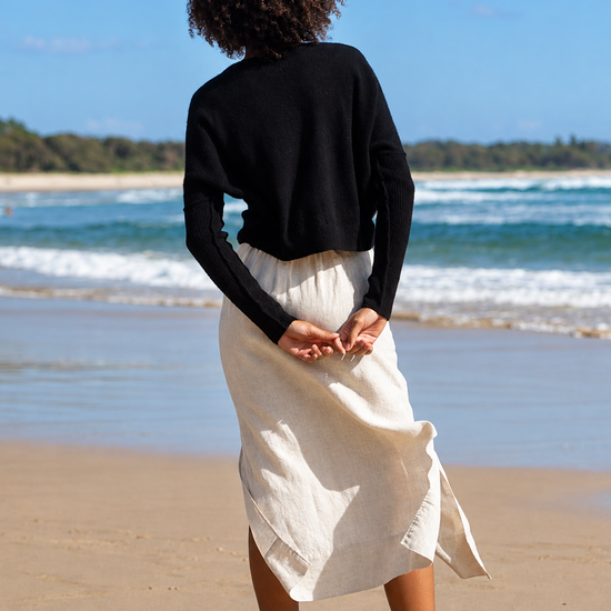 A woman stands on a sandy beach with the ocean and blue sky in the background, viewed from behind. She is wearing a black long-sleeve cropped top and a cream-colored linen midi skirt.