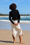 A woman stands on a sandy beach with the ocean and blue sky in the background, viewed from behind. She is wearing a black long-sleeve cropped top and a cream-colored linen midi skirt.