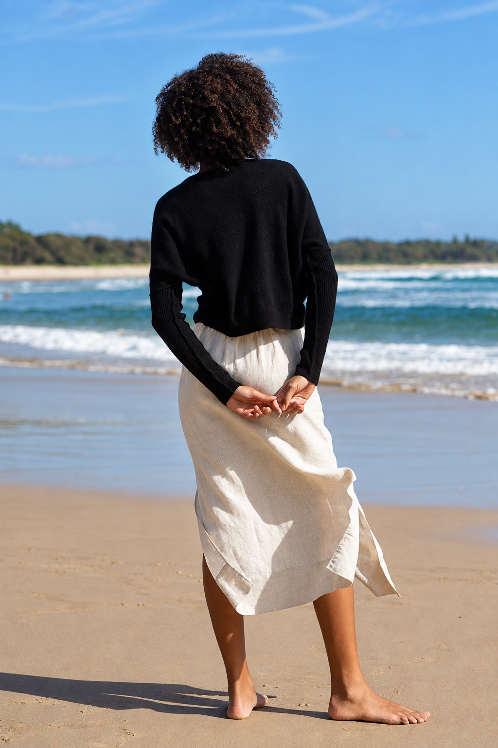A woman stands on a sandy beach with the ocean and blue sky in the background, viewed from behind. She is wearing a black long-sleeve cropped top and a cream-colored linen midi skirt.