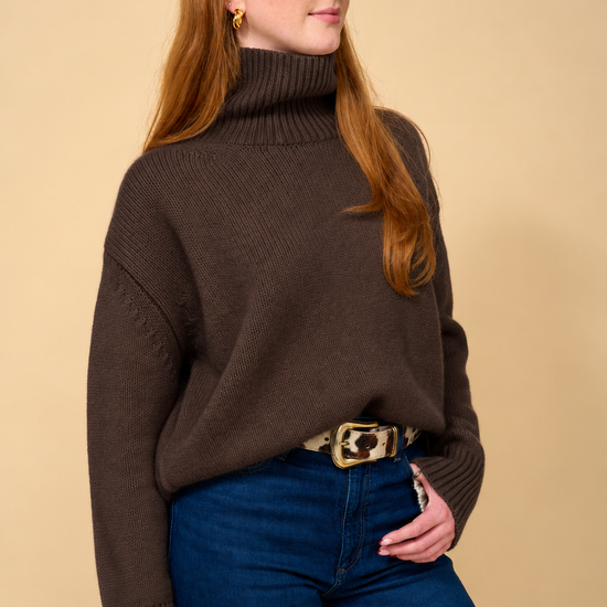 A woman with red hair wearing a dark brown ribbed turtleneck sweater, blue jeans, and a patterned belt, standing against a solid beige background.