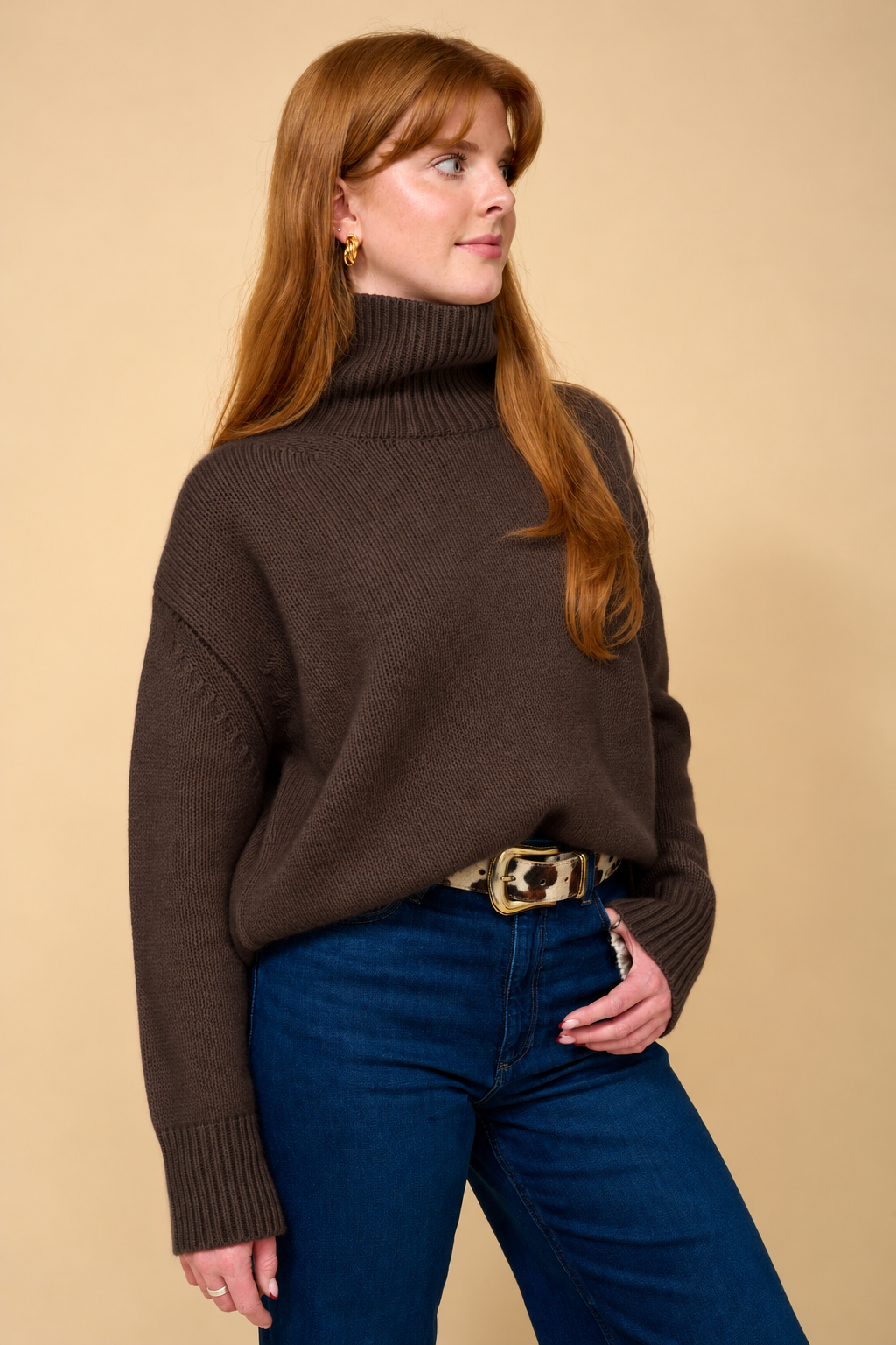 A woman with red hair wearing a dark brown ribbed turtleneck sweater, blue jeans, and a patterned belt, standing against a solid beige background.
