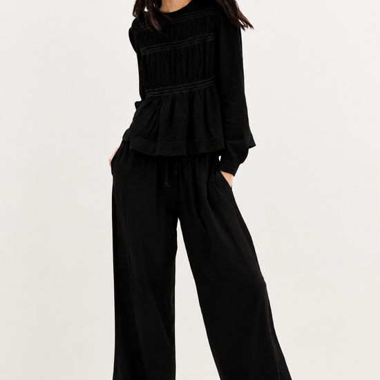 A model wearing a black long-sleeve peplum top, matching black wide-leg trousers, and a black baseball cap, standing against a plain white studio background.