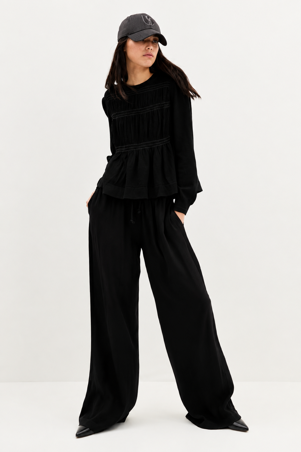 A model wearing a black long-sleeve peplum top, matching black wide-leg trousers, and a black baseball cap, standing against a plain white studio background.