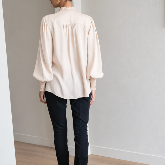 A woman viewed from behind wearing a cream-colored long-sleeve blouse with lace cuffs, black pants, and black knee-high boots in a minimalist room with light gray walls and wood floors.