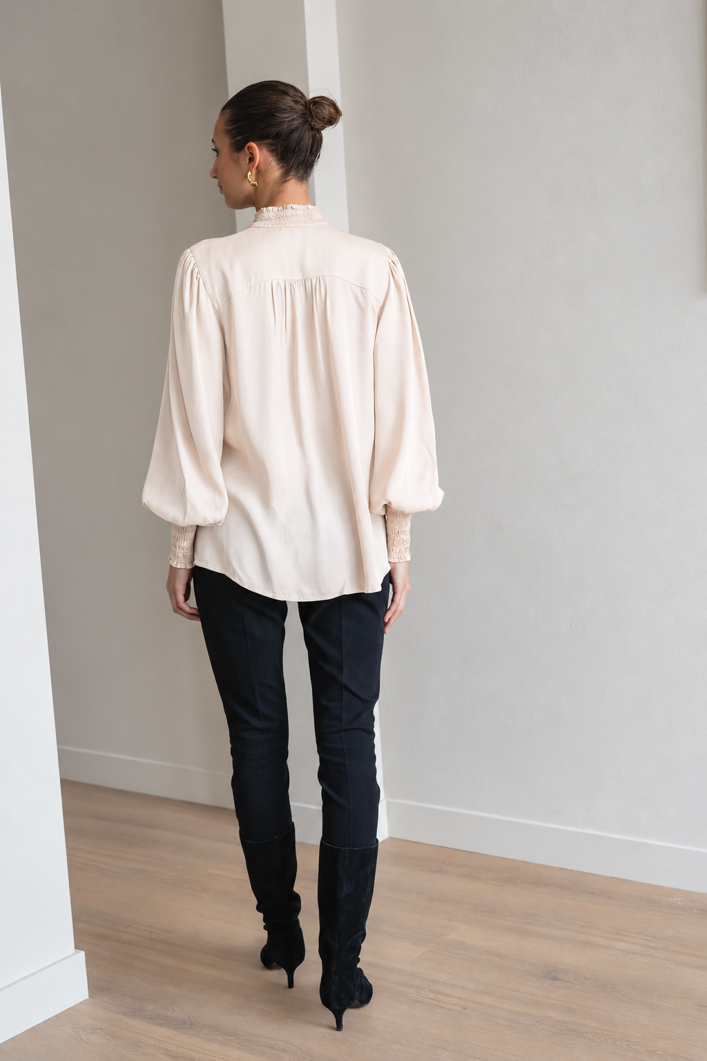 A woman viewed from behind wearing a cream-colored long-sleeve blouse with lace cuffs, black pants, and black knee-high boots in a minimalist room with light gray walls and wood floors.