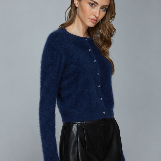 A woman modeling a fuzzy long-sleeve cardigan sweater in deep navy blue, paired with black leather pants, against a solid gray studio background.