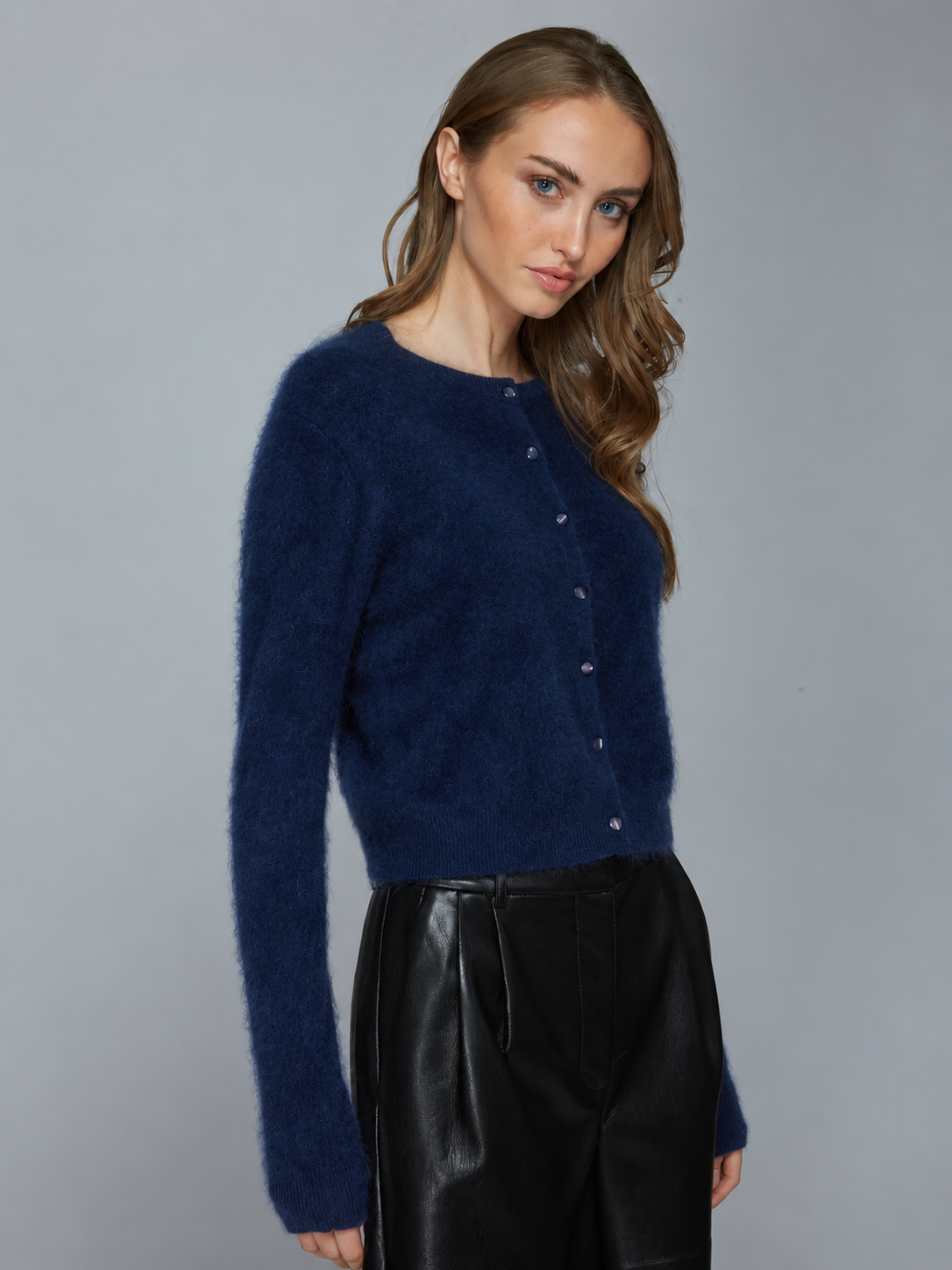 A woman modeling a fuzzy long-sleeve cardigan sweater in deep navy blue, paired with black leather pants, against a solid gray studio background.