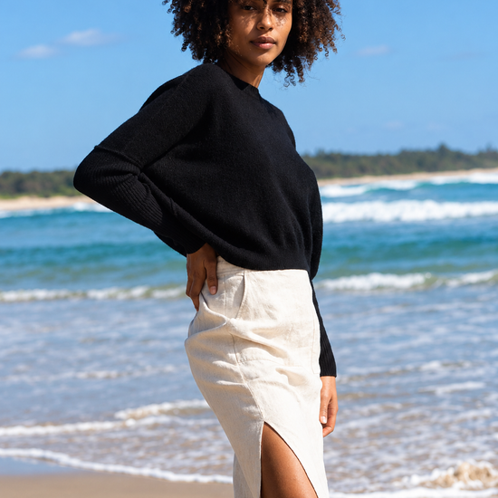A woman stands on a sandy beach wearing a black long-sleeve knit top and a cream high-waisted midi skirt with a thigh-high slit. The background shows the blue ocean and sky.