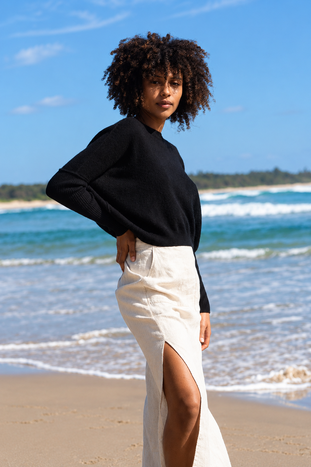 A woman stands on a sandy beach wearing a black long-sleeve knit top and a cream high-waisted midi skirt with a thigh-high slit. The background shows the blue ocean and sky.