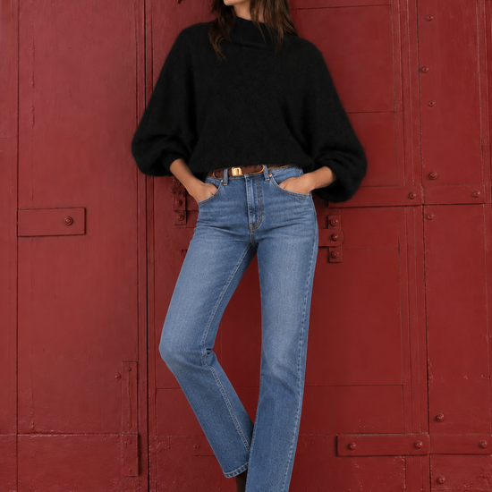 A woman stands in front of a large red metal door wearing a black oversized sweater, light blue straight-leg jeans, and brown ankle boots.