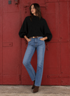 A woman stands in front of a large red metal door wearing a black oversized sweater, light blue straight-leg jeans, and brown ankle boots.