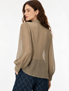 A woman shown from the back wearing a loose, semi-sheer long-sleeve blouse in a taupe-beige color, paired with dark blue patterned trousers, against a plain white studio background.