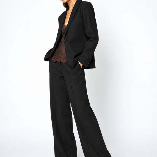 A woman modeling a black tailored suit consisting of a single-breasted blazer and matching wide-leg trousers, styled with a brown lace top and brown pointed-toe heels against a plain light gray studio background.
