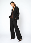 A woman modeling a black tailored suit consisting of a single-breasted blazer and matching wide-leg trousers, styled with a brown lace top and brown pointed-toe heels against a plain light gray studio background.