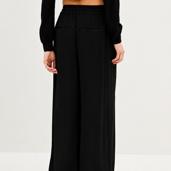 A model from the back wearing black wide-leg trousers with side pockets and a black long-sleeve cropped top, photographed against a plain white studio background.