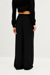 A model from the back wearing black wide-leg trousers with side pockets and a black long-sleeve cropped top, photographed against a plain white studio background.