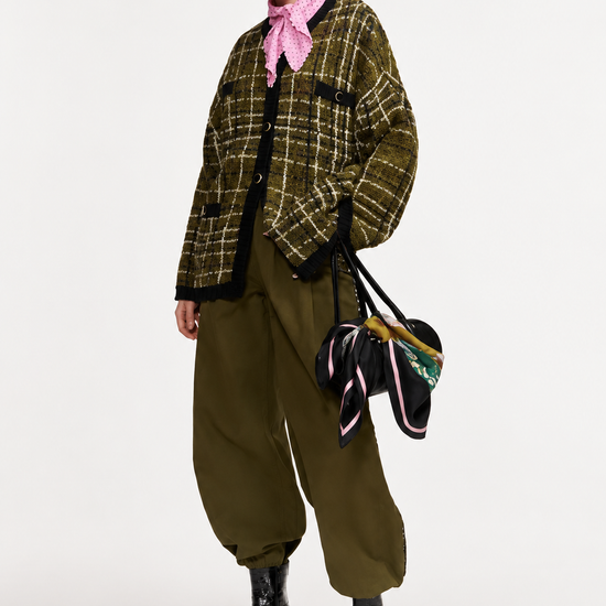 A model wearing a green and black plaid cropped jacket, olive green baggy pants, a pink neck scarf, black sunglasses, and black patent leather boots, holding a black handbag with colorful ribbons.