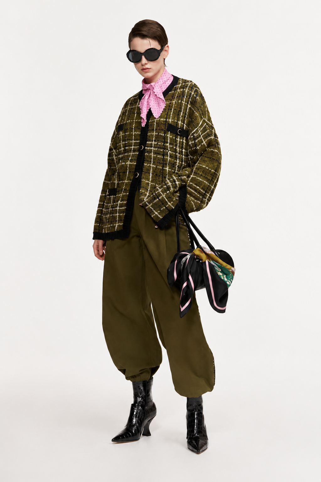 A model wearing a green and black plaid cropped jacket, olive green baggy pants, a pink neck scarf, black sunglasses, and black patent leather boots, holding a black handbag with colorful ribbons.