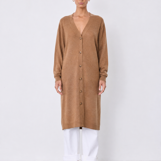 A woman stands against a plain light gray studio wall wearing a long tan button-front cardigan or tunic with long sleeves, white cropped pants, and flat sandals.