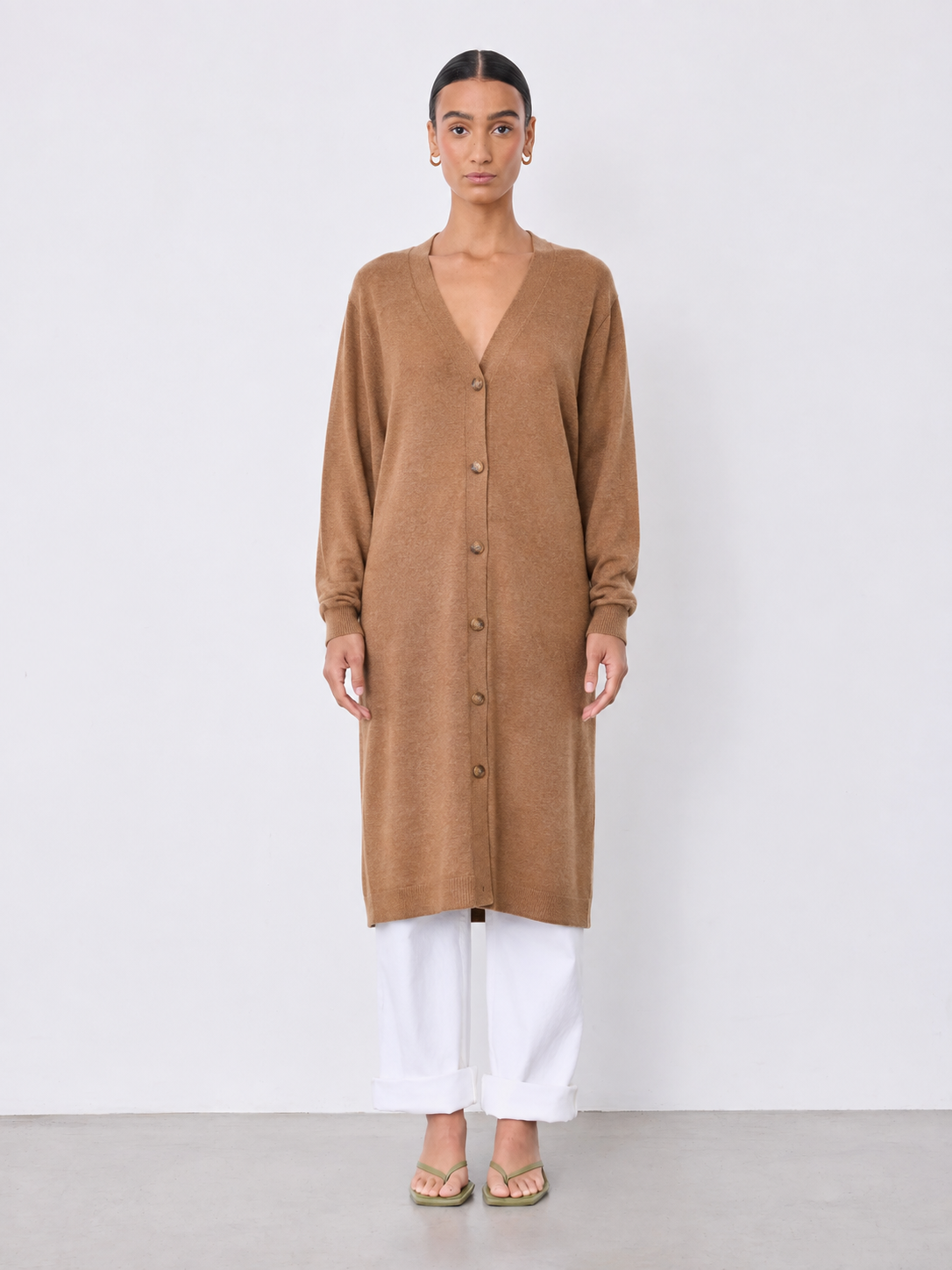 A woman stands against a plain light gray studio wall wearing a long tan button-front cardigan or tunic with long sleeves, white cropped pants, and flat sandals.