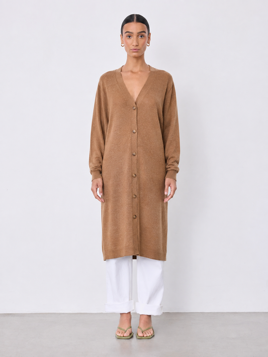 A woman stands against a plain light gray studio wall wearing a long tan button-front cardigan or tunic with long sleeves, white cropped pants, and flat sandals.