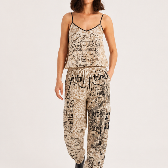 A woman modeling a matching two-piece loungewear set featuring a beige and black newspaper print. The set includes a spaghetti-strap camisole top and matching drawstring pants, styled with black pointed-toe boots against a plain light gray studio background.