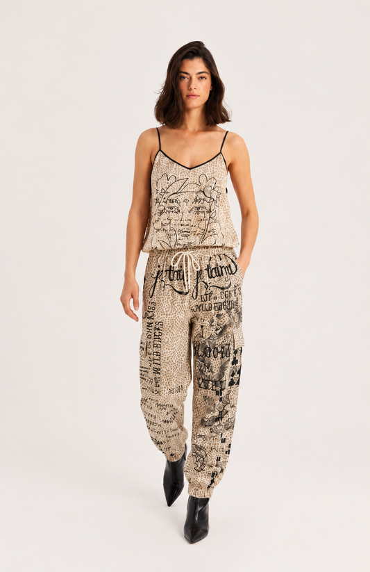 A woman modeling a matching two-piece loungewear set featuring a beige and black newspaper print. The set includes a spaghetti-strap camisole top and matching drawstring pants, styled with black pointed-toe boots against a plain light gray studio background.