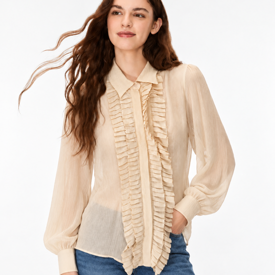 A woman wearing a light beige long-sleeve blouse with a ruffled front placket and collar, paired with blue jeans, photographed against a plain white studio background.