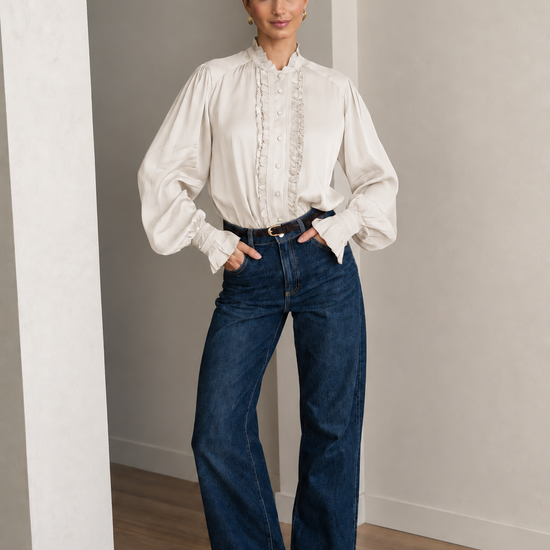 A woman stands in a minimalist room with light gray walls and wood flooring, wearing a cream-colored long-sleeve blouse with lace detailing down the front and dark blue wide-leg jeans.