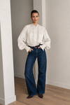 A woman stands in a minimalist room with light gray walls and wood flooring, wearing a cream-colored long-sleeve blouse with lace detailing down the front and dark blue wide-leg jeans.
