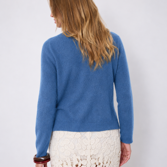 A woman shown from the back wearing a blue knit sweater and a cream lace skirt against a plain light gray studio background.