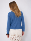 A woman shown from the back wearing a blue knit sweater and a cream lace skirt against a plain light gray studio background.