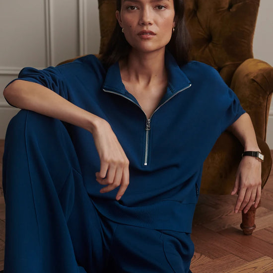A woman wearing a dark blue half-zip jumpsuit sits in a brown velvet armchair in a room with wood floors and white paneled walls.