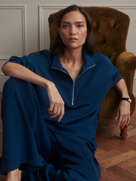 A woman wearing a dark blue half-zip jumpsuit sits in a brown velvet armchair in a room with wood floors and white paneled walls.