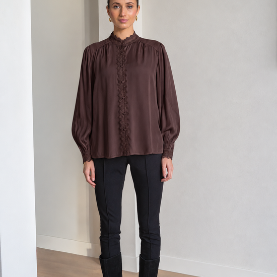 A woman modeling a dark brown long-sleeve blouse with a high gathered neckline and lace-up front, paired with black pants and black knee-high boots in a minimalist indoor setting.
