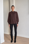 A woman modeling a dark brown long-sleeve blouse with a high gathered neckline and lace-up front, paired with black pants and black knee-high boots in a minimalist indoor setting.