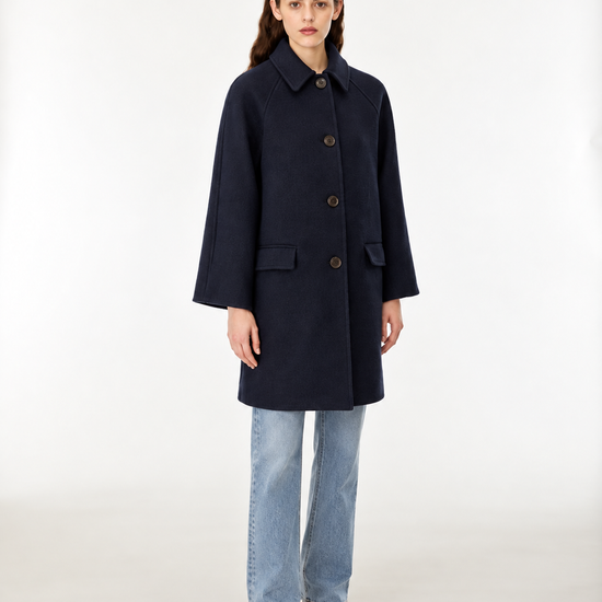 A woman modeling a dark navy blue wool-blend coat with front buttons, paired with light blue jeans and brown shoes, standing against a plain white studio background.